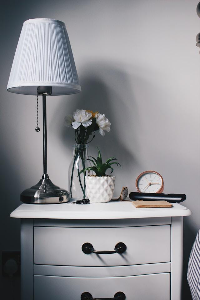 table de chevet blanche, poignées en métal noir, 2 tiroirs avec lampe, fleurs, réveil et télécommande sur la tablette