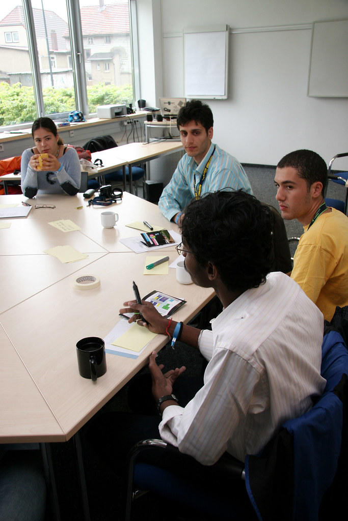 A group of four people sitting in a semi circle of desks, with one person talking and the three others looking at the person talking and listening